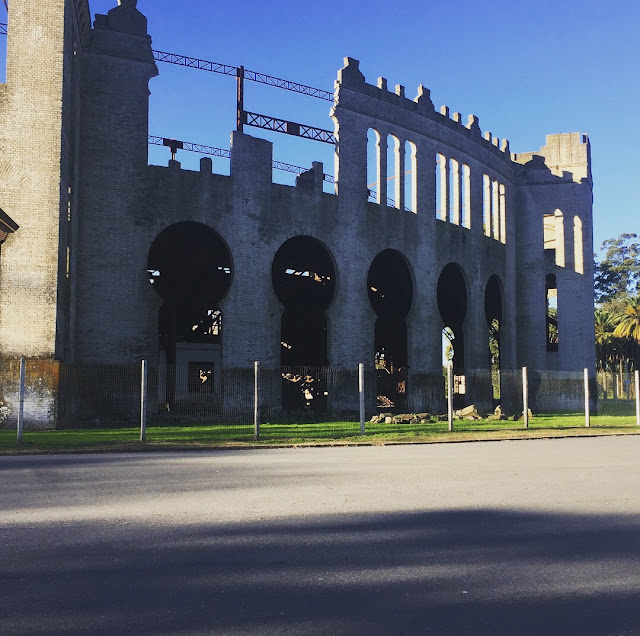 Fachada de uma construção de pedra em ruínas com arcos abertos e ameias em estilo de castelo contra um céu azul limpo, em tons neutros e terrosos.